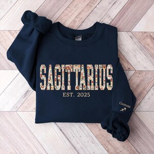 May include: Navy blue sweatshirt with a floral Sagittarius design and the text "EST. 2025". The sweatshirt has a custom Sagittarius symbol embroidered on the sleeve.