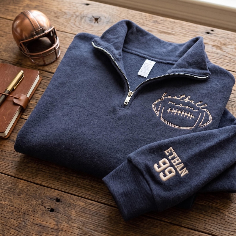 May include: A navy blue quarter-zip sweatshirt featuring a metallic copper-colored embroidered football graphic with the text "football mom" on the chest and the name "ETHAN" above the number "99" embroidered in copper on the sleeve cuff.