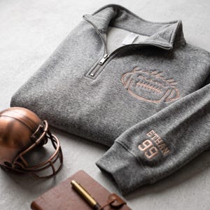 May include: A heather gray quarter-zip pullover featuring copper-colored embroidery of a football and the text football mom on the chest, with the name ETHAN and the number 99 embroidered in copper on the sleeve cuff, near a copper football helmet.