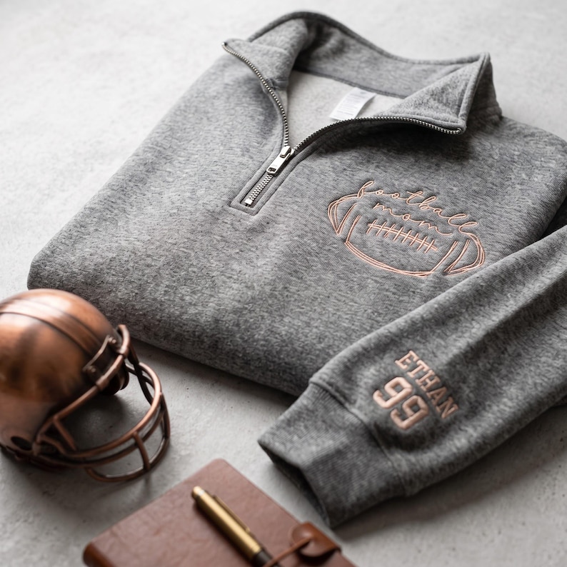 May include: A heather gray quarter-zip pullover featuring copper-colored embroidery of a football and the text football mom on the chest, with the name ETHAN and the number 99 embroidered in copper on the sleeve cuff, near a copper football helmet.