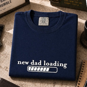 May include: Navy blue t-shirt with the text "new dad loading" in white, along with a loading bar graphic. The shirt is folded neatly, with a small tag visible at the neckline. The shirt is made of a soft material.