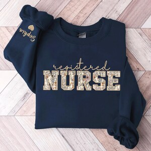 May include: Navy blue sweatshirt with the text "registered NURSE" in white letters with a floral pattern. The text is in a varsity style font.
