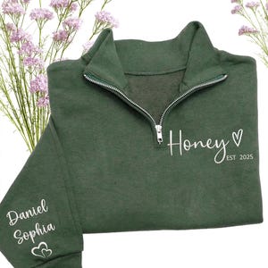 May include: A dark green pullover sweatshirt with a quarter-zip neckline. The word "Honey" and a heart are printed on the front, with "EST 2025" below. The sleeve features the names "Daniel Sophia" and two hearts.