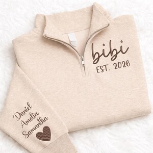 May include: Beige quarter-zip sweatshirt with the word "bibi" and "EST. 2026" embroidered in brown. The left sleeve features the names "Daniel Amelia Samantha" and a brown heart. The sweatshirt has a ribbed collar and cuffs.