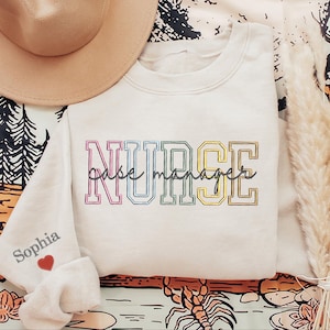 May include: A white sweatshirt with the word "NURSE" embroidered in rainbow colors. The text "take manage" is written in cursive below the word "NURSE". The sweatshirt has a small red heart and the name "Sophia" embroidered on the left sleeve.