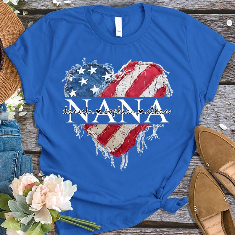 K&ouml;nnte beinhalten: Ein blaues T-Shirt mit einem herzf&ouml;rmigen Design in Form der amerikanischen Flagge. Das Wort "NANA" ist wei&szlig; in der Mitte aufgedruckt, darunter die Namen "daniela sophia ethan". Das T-Shirt ist am unteren Ende gefaltet und gebunden.