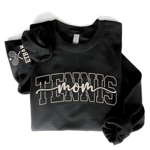 May include: Black long-sleeved sweatshirt featuring the text "TENNIS mom" in white block and script lettering. The left sleeve displays "ETHAN" and a tennis racket graphic. The sweatshirt is folded on a white background.