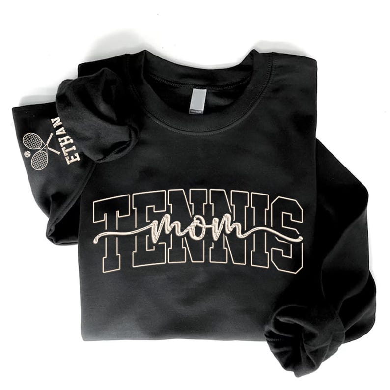 May include: Black long-sleeved sweatshirt featuring the text "TENNIS mom" in white block and script lettering. The left sleeve displays "ETHAN" and a tennis racket graphic. The sweatshirt is folded on a white background.