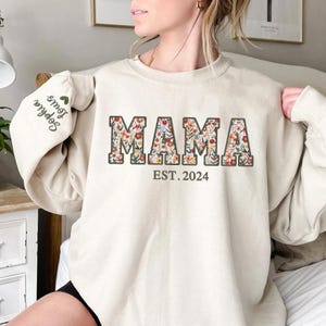 May include: Beige sweatshirt with the word "MAMA" in floral print, outlined in brown, and the text "EST. 2024" below. The left sleeve has the embroidered names "Sophia Louis" with a green heart. A casual, comfortable top.