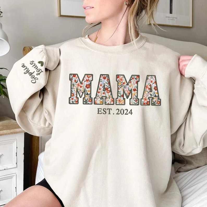 May include: Beige sweatshirt with the word "MAMA" in floral print, outlined in brown, and the text "EST. 2024" below. The left sleeve has the embroidered names "Sophia Louis" with a green heart. A casual, comfortable top.
