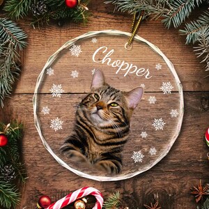 May include: A clear glass Christmas ornament with a tabby cat image and the name "Chopper" printed on it. The ornament is decorated with snowflake designs.