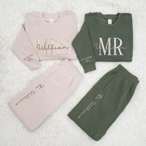 May include: Matching beige and olive green loungewear sets. Each set includes a crewneck sweatshirt and sweatpants. The sweatshirts are embroidered with "MRS" and "MR" with the last name "William" in gold lettering. The date "09.19.2025" is on the sleeves.