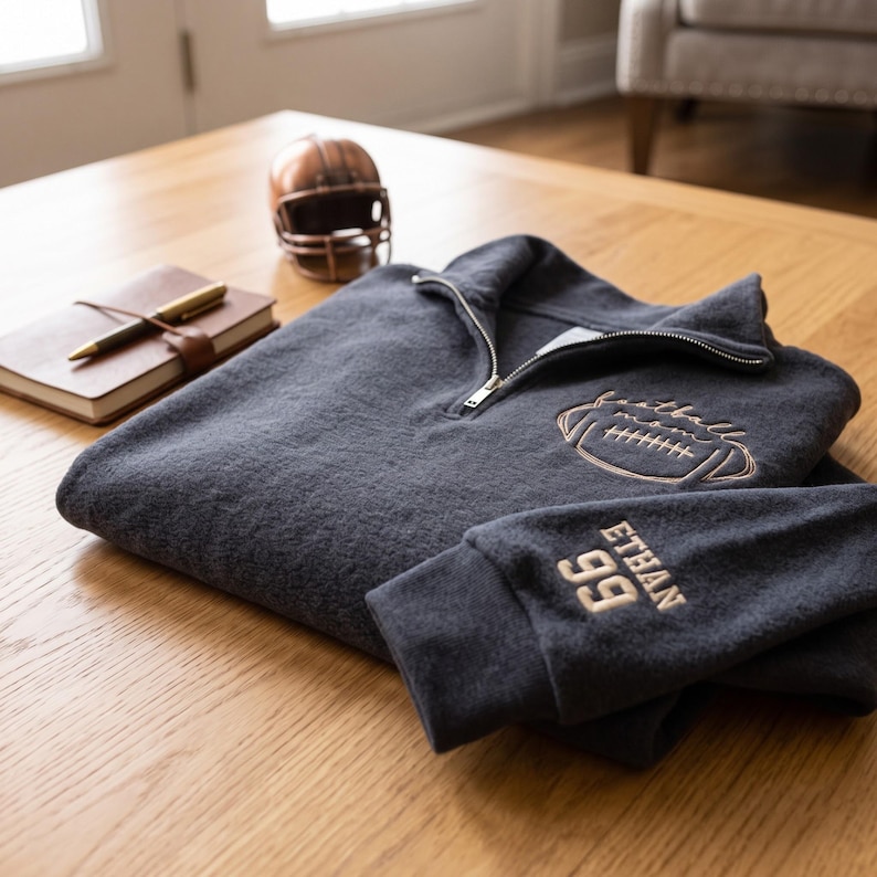 May include: A dark charcoal gray quarter-zip pullover featuring an embroidered football graphic on the chest with the text football mom and the name ETHAN with the number 99 embroidered in light tan thread on the sleeve cuff.