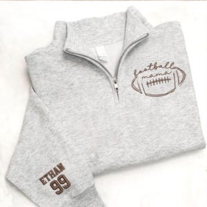 May include: A light gray quarter-zip sweatshirt featuring a brown embroidered football graphic with the text "football mama" on the chest and the name "ETHAN" above the number "99" in brown embroidery on the sleeve cuff.