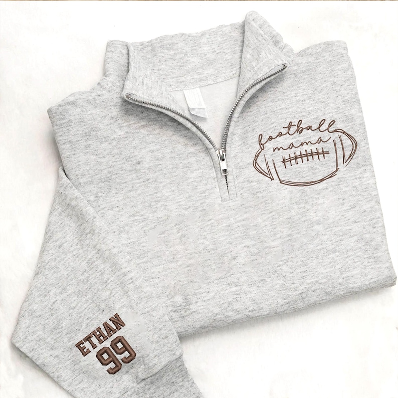 May include: A light gray quarter-zip sweatshirt featuring a brown embroidered football graphic with the text "football mama" on the chest and the name "ETHAN" above the number "99" in brown embroidery on the sleeve cuff.