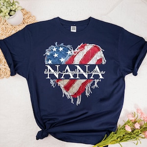 K&ouml;nnte beinhalten: Marineblaues T-Shirt mit einem herzf&ouml;rmigen Design der amerikanischen Flagge. Das Wort "NANA" ist in der Mitte des Herzens platziert, darunter die Namen "Daniel, Sophia, Ethan". Das Design hat einen Used-Look.