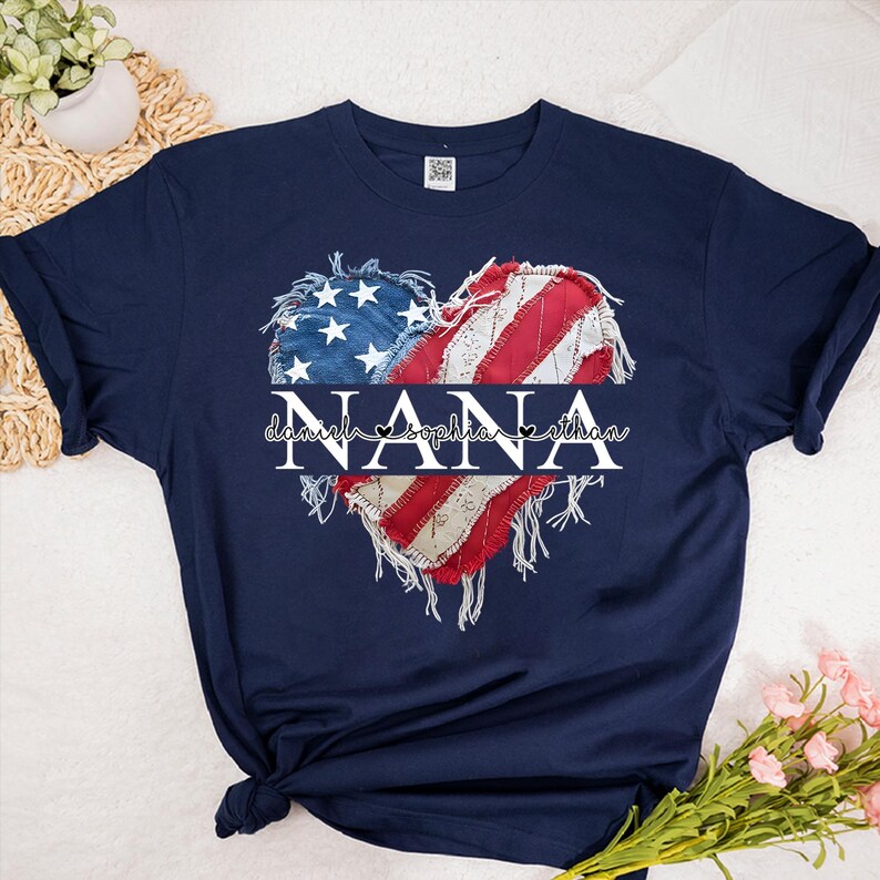 K&ouml;nnte beinhalten: Marineblaues T-Shirt mit einem herzf&ouml;rmigen Design der amerikanischen Flagge. Das Wort "NANA" ist in der Mitte des Herzens platziert, darunter die Namen "Daniel, Sophia, Ethan". Das Design hat einen Used-Look.