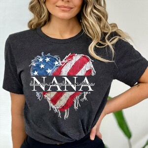 K&ouml;nnte beinhalten: Dunkelgraues T-Shirt mit einem herzf&ouml;rmigen Design der amerikanischen Flagge. Das Herz besteht aus blauem, wei&szlig;em und rotem Stoff mit einem Used-Look. Das Wort "NANA" ist in Wei&szlig; gedruckt, darunter die Namen "Daniel", "Sophia" und "Ethan".