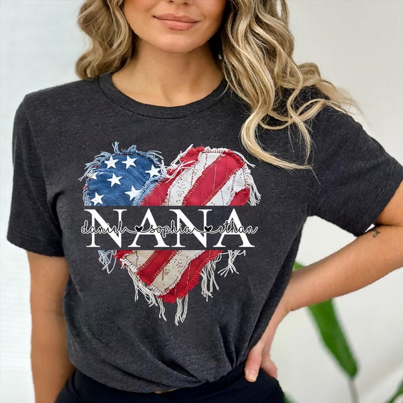 K&ouml;nnte beinhalten: Dunkelgraues T-Shirt mit einem herzf&ouml;rmigen Design der amerikanischen Flagge. Das Herz besteht aus blauem, wei&szlig;em und rotem Stoff mit einem Used-Look. Das Wort "NANA" ist in Wei&szlig; gedruckt, darunter die Namen "Daniel", "Sophia" und "Ethan".