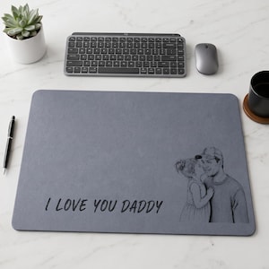 May include: A gray rectangular desk mat with the text "I LOVE YOU DADDY" and a black and white illustration of a father and child. A keyboard, mouse, pen, succulent, and mug are also visible.