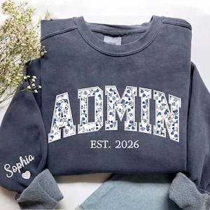 May include: A dark gray sweatshirt with the word "ADMIN" in white, floral-patterned letters. The text "EST. 2026" is below the word "ADMIN". The name "Sophia" is embroidered on the sleeve.