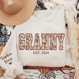 May include: A cream-colored sweatshirt with the word "Granny" in brown letters with a floral pattern. The text "EST. 2024" is below the word "Granny".
