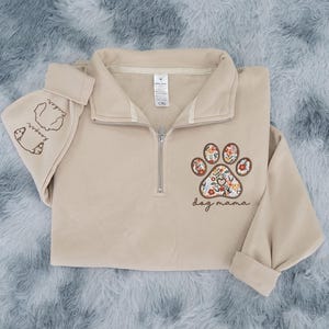 May include: A beige quarter-zip sweatshirt with a floral paw print design and the words "dog mama." The left sleeve has the words "Tucker keeper" and a dog illustration. The sweatshirt is folded on a soft, textured surface.