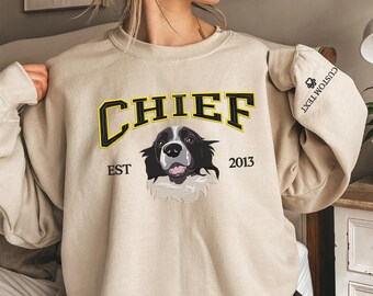 May include: Beige sweatshirt with the word "CHIEF" in black and yellow lettering above a black and white dog graphic. The text "EST 2013" is below the dog. The right sleeve has the text "CUSTOM TEXT" and a paw print.