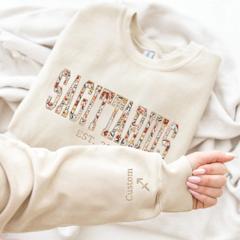May include: A beige sweatshirt with a floral Sagittarius design and the text "EST. 20..." on the front. The sleeve has a small Sagittarius symbol and the word "Custom".