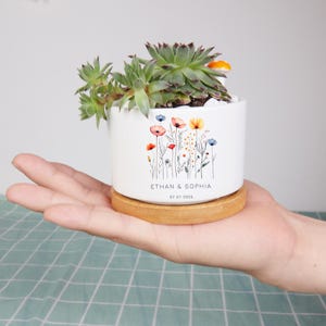 May include: A white ceramic planter with a floral design and the text "ETHAN & SOPHIA 07.07.2025" holds a succulent plant. The planter sits on a round wooden base. The succulent has green leaves and is filled with white stones.