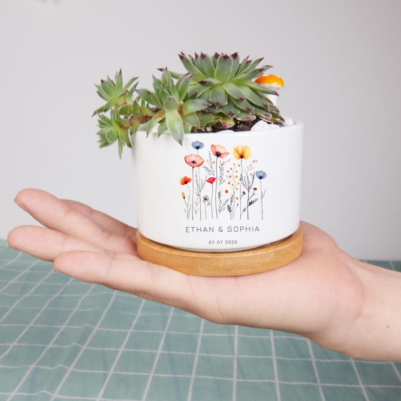 May include: A white ceramic planter with a floral design and the text "ETHAN & SOPHIA 07.07.2025" holds a succulent plant. The planter sits on a round wooden base. The succulent has green leaves and is filled with white stones.