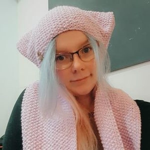 May include: A pink knitted hat with cat ears and a matching scarf. The hat has a button detail on the front.