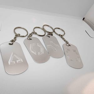 Könnte beinhalten: Vier silberne Metall-Schlüsselanhänger mit eingravierten geometrischen Designs. Jeder Schlüsselanhänger hat ein rechteckiges Etikett mit einem anderen Symbol, das an einem Metallring und einer Kette befestigt ist. Die Designs umfassen einen Pfeil nach oben, ein Sechseck, ein Kreuz und ein Quadrat.