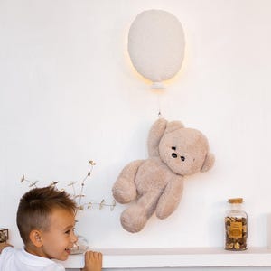 Könnte beinhalten: Eine skurrile Szene im Kinderzimmer mit einem beigen Teddybären, der an einer ballonförmigen Leuchte befestigt ist. Ein Kind in einem weißen Hemd und einer braunen Hose steht in der Nähe einer Wiege aus Korbgeflecht mit Kissen. Der Kamin ist aus weißen Ziegeln.