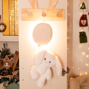 Pode incluir: Um cabide de madeira com detalhes de orelhas de coelho e tr&ecirc;s pinos, montado em uma porta branca. Abaixo, um coelho de pel&uacute;cia branco e fofo pendura, com uma luz em forma de bal&atilde;o brilhante. Decora&ccedil;&otilde;es de f&eacute;rias.