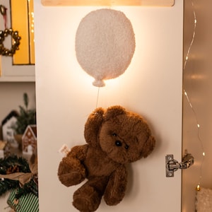 Pode incluir: Um cabide de parede de madeira com tr&ecirc;s pinos e orelhas de coelho. Abaixo, uma lumin&aacute;ria branca em forma de bal&atilde;o ilumina um urso de pel&uacute;cia marrom pendurado por um fio. A cena &eacute; montada em uma porta branca.