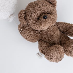 Pode incluir: Um urso de pel&uacute;cia marrom com uma textura fofa, segurando um bal&atilde;o branco. O urso tem olhos pretos e uma etiqueta que diz "ZAYKO". O brinquedo est&aacute; em um fundo branco.
