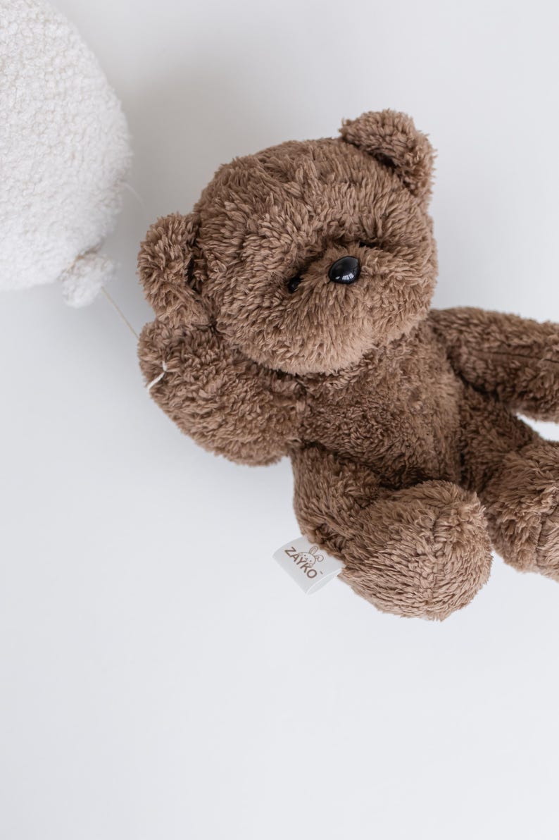 Pode incluir: Um urso de pel&uacute;cia marrom com uma textura fofa, segurando um bal&atilde;o branco. O urso tem olhos pretos e uma etiqueta que diz "ZAYKO". O brinquedo est&aacute; em um fundo branco.