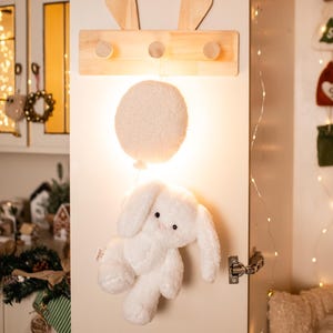 Pode incluir: Um brinquedo de coelho de pel&uacute;cia branco com orelhas compridas e um corpo fofo pendura-se sob uma luz redonda branca em forma de bal&atilde;o. Acima, um cabide de madeira com recortes de orelhas de coelho e tr&ecirc;s pinos &eacute; montado em uma porta branca.