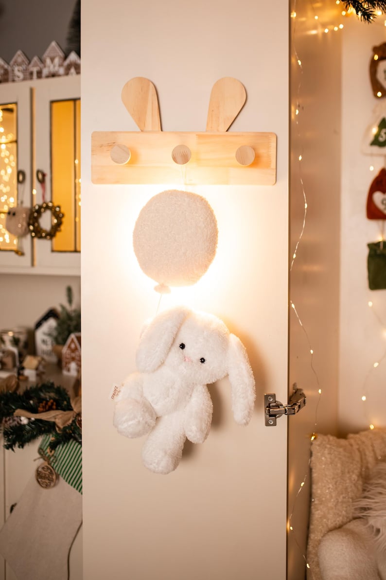 Pode incluir: Um brinquedo de coelho de pel&uacute;cia branco com orelhas compridas e um corpo fofo pendura-se sob uma luz redonda branca em forma de bal&atilde;o. Acima, um cabide de madeira com recortes de orelhas de coelho e tr&ecirc;s pinos &eacute; montado em uma porta branca.