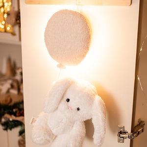 Pode incluir: Um brinquedo de coelho branco de pel&uacute;cia com orelhas compridas e um nariz rosa pendura-se sob uma luz redonda e fofa em forma de bal&atilde;o branco. A luz e o coelho est&atilde;o presos a uma decora&ccedil;&atilde;o de parede de madeira com tr&ecirc;s pinos redondos.