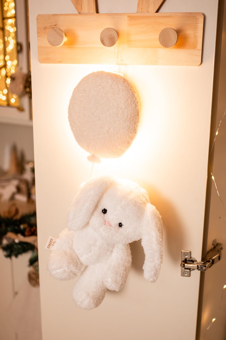 Pode incluir: Um brinquedo de coelho branco de pel&uacute;cia com orelhas compridas e um nariz rosa pendura-se sob uma luz redonda e fofa em forma de bal&atilde;o branco. A luz e o coelho est&atilde;o presos a uma decora&ccedil;&atilde;o de parede de madeira com tr&ecirc;s pinos redondos.