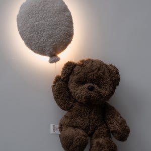 Pode incluir: Um urso de pel&uacute;cia marrom segurando uma lumin&aacute;ria branca e fofa em forma de bal&atilde;o. A lumin&aacute;ria est&aacute; iluminada, emitindo um brilho quente. O urso tem uma etiqueta anexada. O fundo &eacute; uma parede branca lisa.