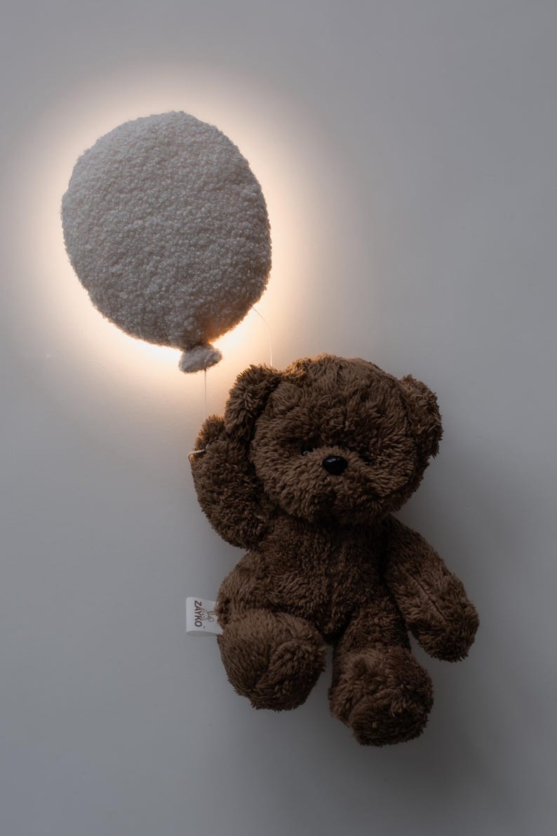 Pode incluir: Um urso de pel&uacute;cia marrom segurando uma lumin&aacute;ria branca e fofa em forma de bal&atilde;o. A lumin&aacute;ria est&aacute; iluminada, emitindo um brilho quente. O urso tem uma etiqueta anexada. O fundo &eacute; uma parede branca lisa.