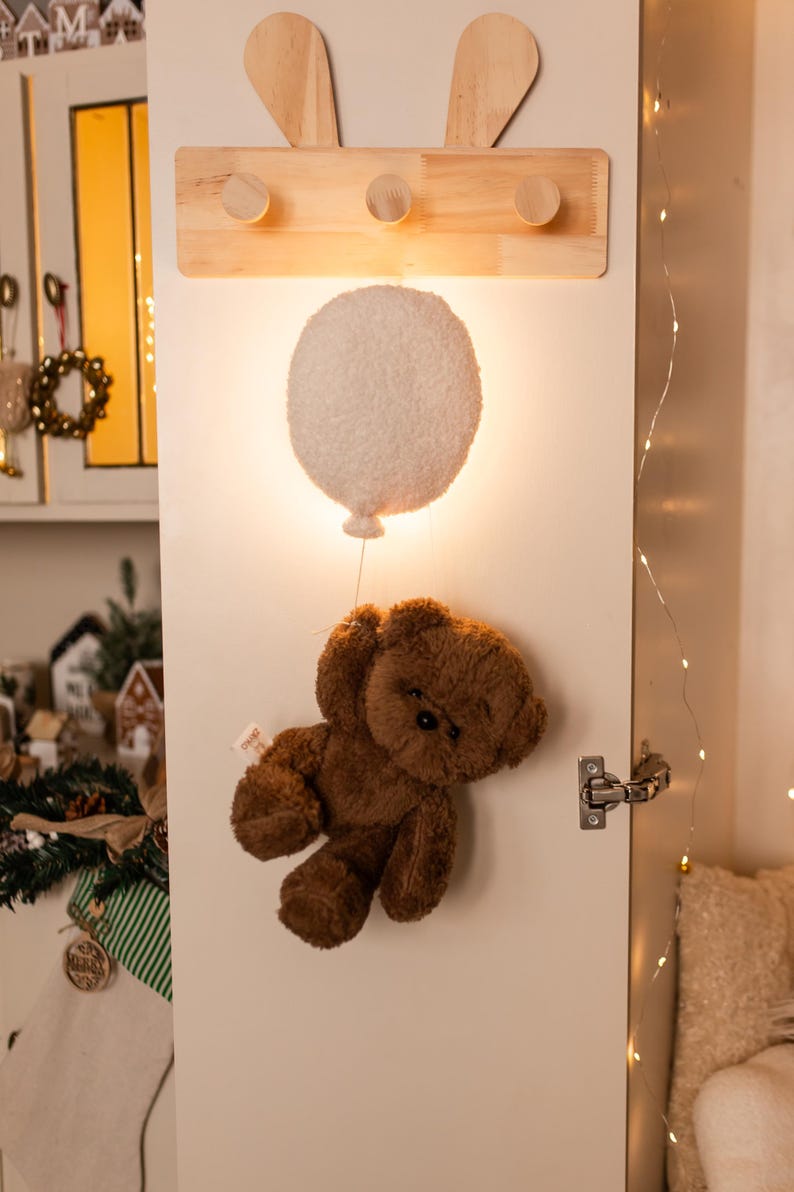 Pode incluir: Um cabide de parede de madeira com tr&ecirc;s pinos e orelhas de coelho. Abaixo, uma lumin&aacute;ria branca em forma de bal&atilde;o ilumina um urso de pel&uacute;cia marrom pendurado por um fio. A cena &eacute; montada em uma porta branca.