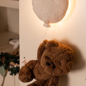 Pode incluir: Um urso de pel&uacute;cia castanho suspenso de uma lumin&aacute;ria em forma de bal&atilde;o branco e fofo. O urso tem um design cl&aacute;ssico com uma apar&ecirc;ncia macia e texturizada. A lumin&aacute;ria de bal&atilde;o &eacute; montada em uma parede branca, criando um elemento decorativo.