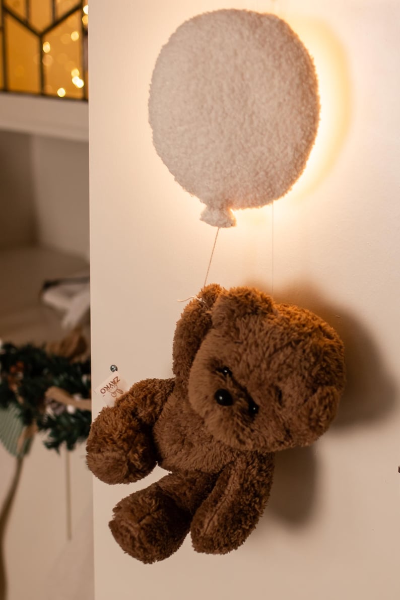 Pode incluir: Um urso de pel&uacute;cia castanho suspenso de uma lumin&aacute;ria em forma de bal&atilde;o branco e fofo. O urso tem um design cl&aacute;ssico com uma apar&ecirc;ncia macia e texturizada. A lumin&aacute;ria de bal&atilde;o &eacute; montada em uma parede branca, criando um elemento decorativo.
