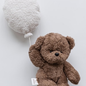 Pode incluir: Um urso de pel&uacute;cia marrom segurando um bal&atilde;o branco. O urso tem um nariz preto e orelhas pequenas. O bal&atilde;o &eacute; redondo e fofo, conectado ao urso por um fio branco fino. Uma etiqueta com o texto "ZAYKO" est&aacute; presa ao urso.