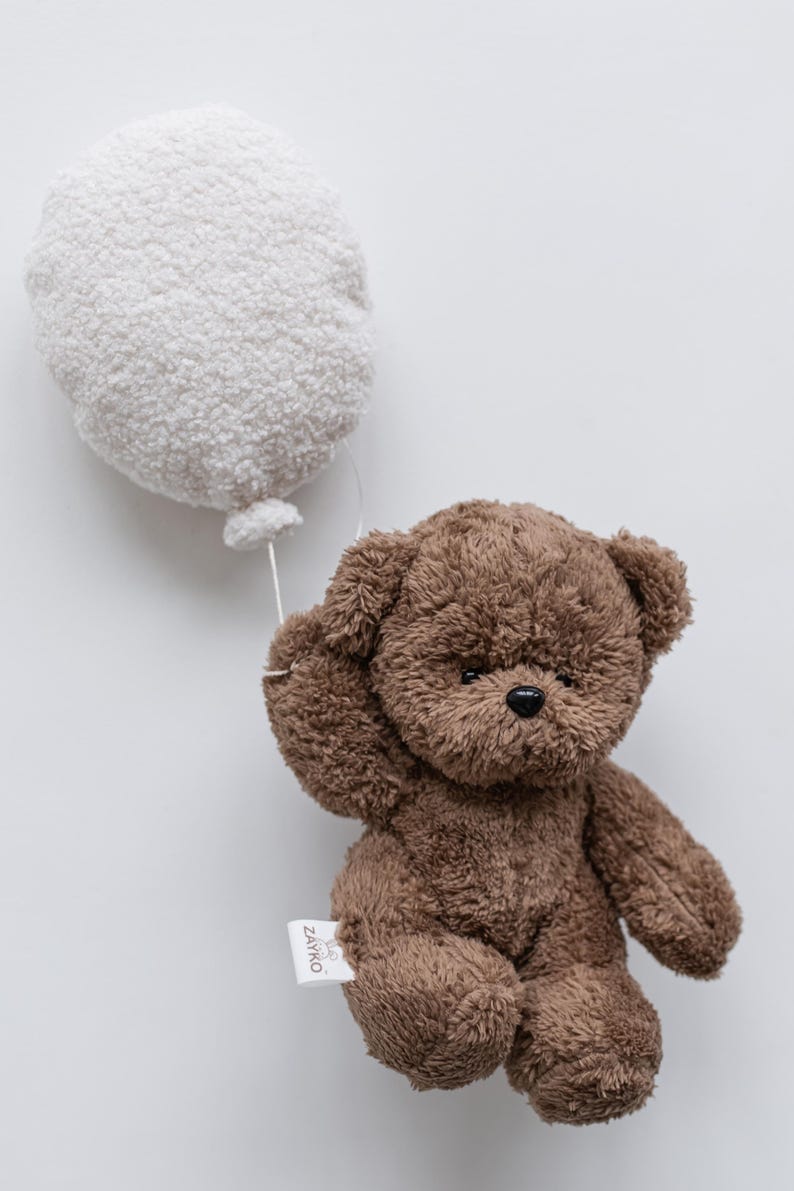 Pode incluir: Um urso de pel&uacute;cia marrom segurando um bal&atilde;o branco. O urso tem um nariz preto e orelhas pequenas. O bal&atilde;o &eacute; redondo e fofo, conectado ao urso por um fio branco fino. Uma etiqueta com o texto "ZAYKO" est&aacute; presa ao urso.