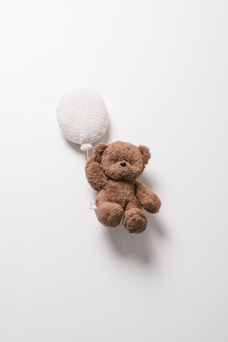 Pode incluir: Um urso de pel&uacute;cia marrom segurando um bal&atilde;o branco e fofo. O urso tem um pequeno nariz preto e &eacute; feito de material macio e texturizado. O bal&atilde;o &eacute; redondo e tamb&eacute;m tem uma apar&ecirc;ncia macia e texturizada. O fundo &eacute; branco liso.